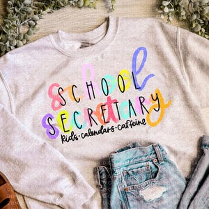 Può includere: Una felpa grigio chiaro con la scritta "SCHOOL SECRETARY" in lettere colorate e giocose. Sotto, le parole "kids • calendars • caffeine". Sono visibili anche dei jeans di denim chiaro.