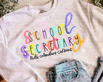 May include: A light gray sweatshirt with the words "SCHOOL SECRETARY" in colorful, playful lettering. Below the main text are the words "kids • calendars • caffeine". The sweatshirt is paired with light wash denim jeans.