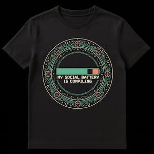 Könnte beinhalten: Schwarzes T-Shirt mit einem kreisförmigen Grafikdesign. Die Grafik zeigt ein grünes Batteriesymbol mit einem Fortschrittsbalken und dem Text "MY SOCIAL BATTERY IS COMPILING". Das Design wird von einem Leiterplatten-ähnlichen Rand mit Binärcode umrahmt.