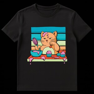 May include: Black t-shirt featuring a cartoon cat baking cupcakes. The cat is orange with a pink frosting swirl on its head and is holding a whisk. A blue bowl with pink batter and cupcakes are on the table.