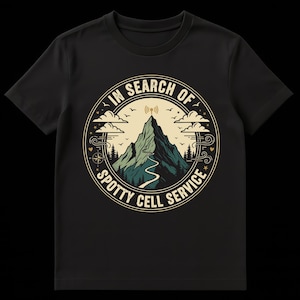 Könnte beinhalten: Schwarzes T-Shirt mit einem kreisförmigen Grafikdesign, das eine Berglandschaft zeigt. Das Design umfasst einen Berggipfel mit einem Mobilfunksymbol, eine kurvenreiche Straße, Bäume, Wolken und den Text "IN SEARCH OF SPOTTY CELL SERVICE."