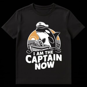 Può includere: T-shirt nera con un'orca dei cartoni animati con un cappello da capitano, una barca e un volante. Il testo "I AM THE CAPTAIN NOW" è bianco. Lo sfondo presenta un tramonto giallo e arancione.