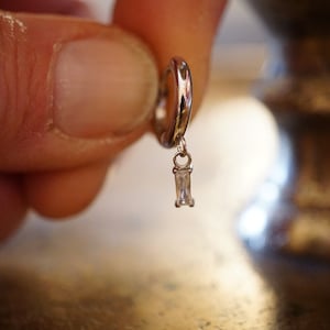 Peut inclure: Une boucle d'oreille en argent avec une pierre transparente rectangulaire pendante.