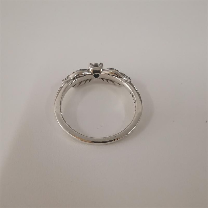 May include: A silver ring with a heart-shaped design and a small black stone in the center. The ring is decorated with intricate details on the sides.