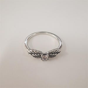 May include: A silver ring with a clear stone in the center and two wing-shaped designs on either side of the stone.