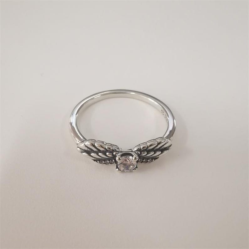 May include: A silver ring with a clear stone in the center and two wing-shaped designs on either side of the stone.