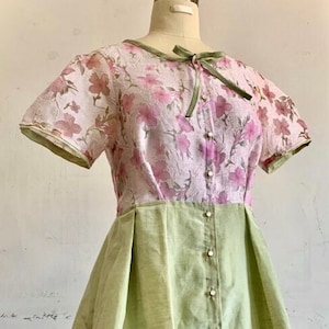 Könnte beinhalten: Ein hellgrünes und rosa Kleid mit Blumendruck. Das Kleid hat einen Knopfleisten vorne und eine Schleife am Ausschnitt.