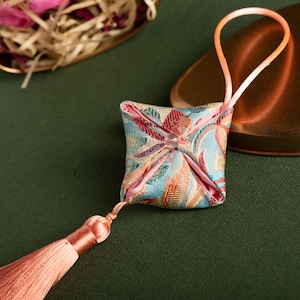May include: A small, square, blue and pink patterned fabric pouch with a pink tassel. The pouch is hanging from a pink ribbon loop.