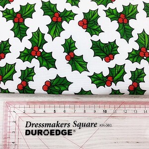 May include: White fabric with a repeating pattern of green holly leaves and red berries. The fabric is measured with a ruler that reads "Dressmakers Square DUR-O-EDGE KR-360".