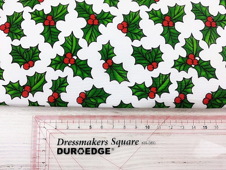 May include: White fabric with a repeating pattern of green holly leaves and red berries. The fabric is measured with a ruler that reads "Dressmakers Square DUR-O-EDGE KR-360".