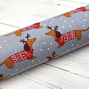 May include: Gray fabric with a repeating pattern of brown dachshunds wearing red and white sweaters with reindeer antlers. The dachshunds are on a white background with white dots.