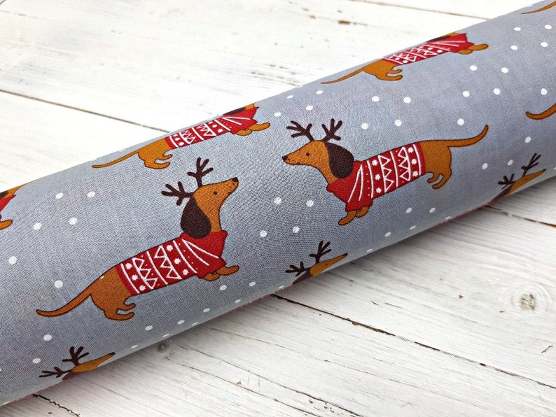 May include: Gray fabric with a repeating pattern of brown dachshunds wearing red and white sweaters with reindeer antlers. The dachshunds are on a white background with white dots.