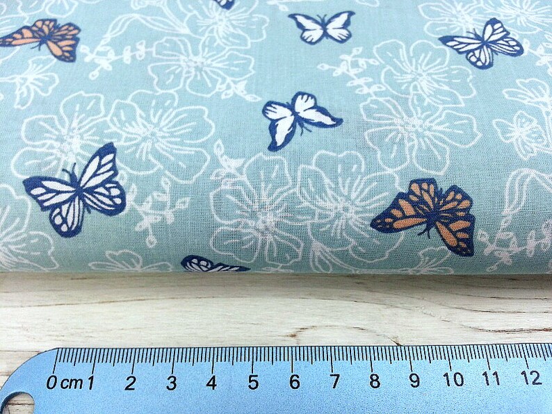 May include: A light blue fabric with a white floral pattern and colorful butterfly designs. The fabric is measured with a ruler in inches.