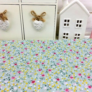 May include: A light blue fabric with a small floral print in pink, yellow, and green. The fabric is shown on a table with a white decorative house and two white drawers with heart-shaped handles.