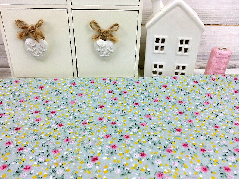 May include: A light blue fabric with a small floral print in pink, yellow, and green. The fabric is shown on a table with a white decorative house and two white drawers with heart-shaped handles.