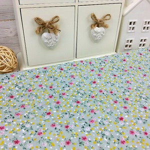 May include: A light blue fabric with a small floral pattern in pink, yellow, and green. The fabric is shown on a table with a white decorative shelf in the background.