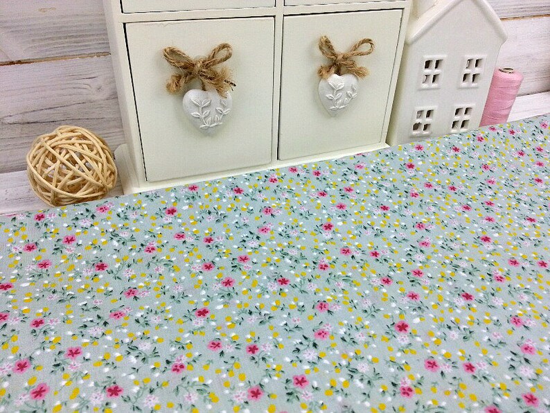 May include: A light blue fabric with a small floral pattern in pink, yellow, and green. The fabric is shown on a table with a white decorative shelf in the background.