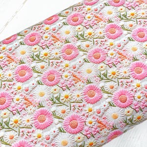Op de afbeelding: Witte stof met een herhalend patroon van roze en witte geborduurde bloemen. De bloemen zijn gestileerd en lijken op madeliefjes.