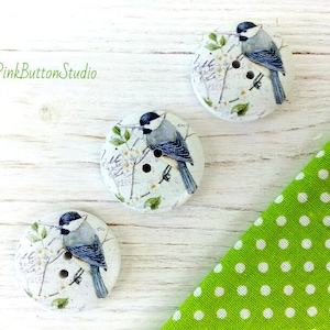 May include: Three white wooden buttons featuring a bluebird design. The buttons have a white background with a floral pattern and a bluebird perched on a branch. The buttons are arranged in a triangular shape on a white wooden surface.