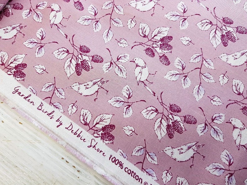 Op de afbeelding: Roze stof met een wit bloemenpatroon met vogels en bessen. De stof heeft de tekst "Garden Birds by Debbie Shore 100% cotton" erop gedrukt.