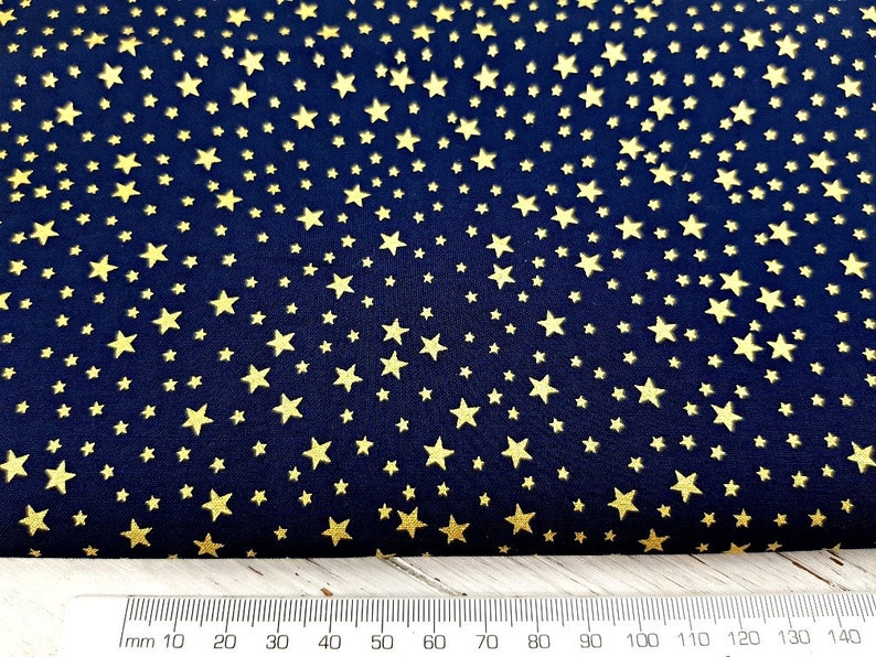 May include: Navy blue fabric with a repeating pattern of gold stars. The stars vary in size and are scattered across the dark blue background. A ruler with millimeter markings is at the bottom of the image.