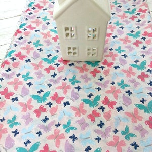 May include: White ceramic house with cut-out windows on a pink fabric with a butterfly pattern. The butterflies are in shades of pink, blue, and green.