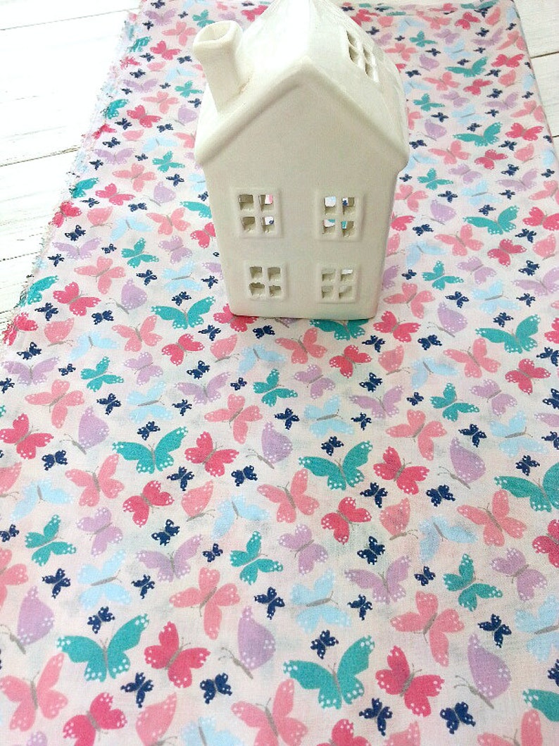 May include: White ceramic house with cut-out windows on a pink fabric with a butterfly pattern. The butterflies are in shades of pink, blue, and green.