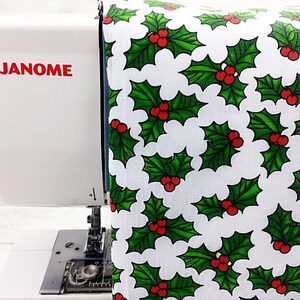 May include: A white sewing machine with the word "JANOME" printed in red on the side. A white fabric with a green holly leaf and red berry pattern is being fed into the machine.