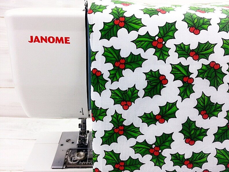 May include: A white sewing machine with the word "JANOME" printed in red on the side. A white fabric with a green holly leaf and red berry pattern is being fed into the machine.