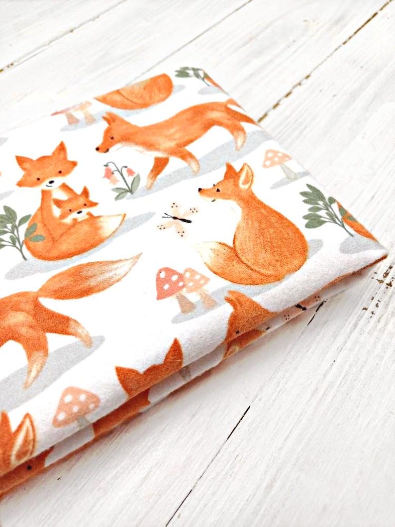 May include: White fabric with a repeating pattern of orange foxes and mushrooms. The foxes are depicted in various poses, including a mother fox carrying a baby fox.