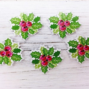 May include: Five wooden Christmas decorations shaped like holly sprigs with red berries. The decorations are green and white with a painted holly leaf and berry design.