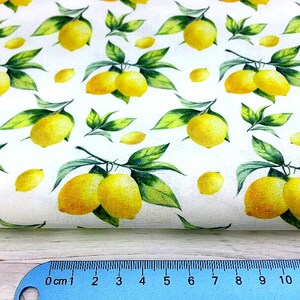 May include: White fabric with a repeating pattern of yellow lemons and green leaves. The lemons are arranged in clusters of two or three, and the leaves are scattered around them.