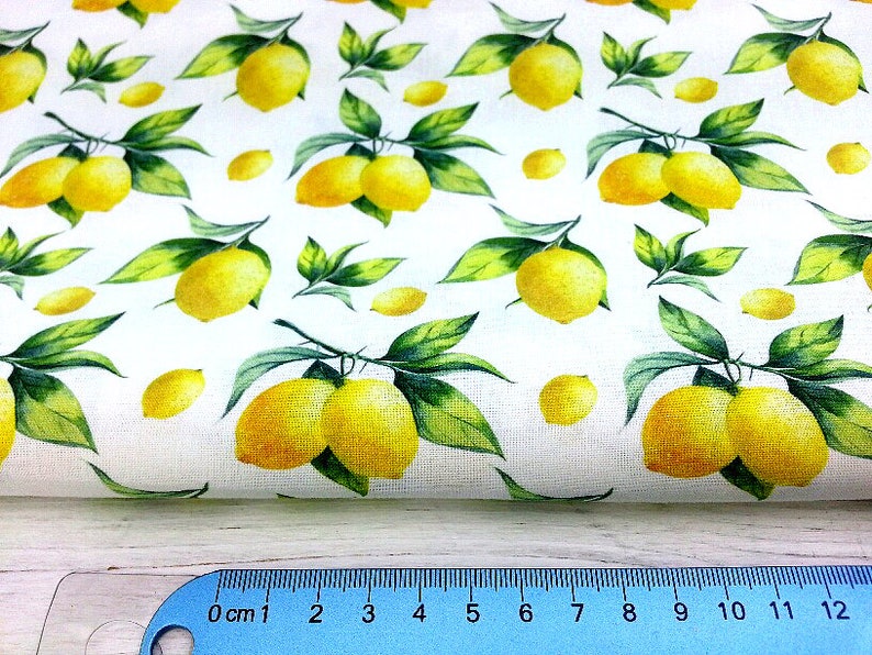 May include: White fabric with a repeating pattern of yellow lemons and green leaves. The lemons are arranged in clusters of two or three, and the leaves are scattered around them.