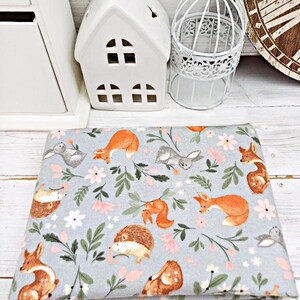 May include: A gray fabric with a repeating pattern of woodland animals and flowers. The animals include foxes, squirrels, hedgehogs, and rabbits. The flowers are pink and white.