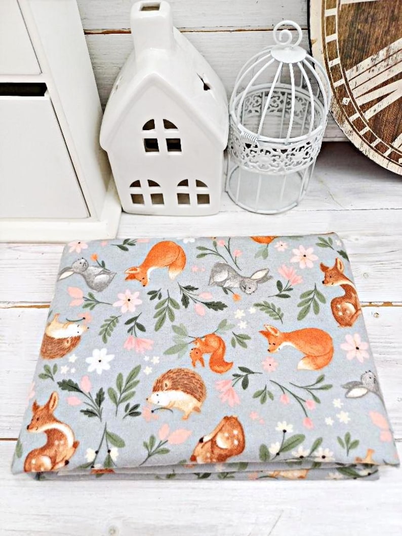 May include: A gray fabric with a repeating pattern of woodland animals and flowers. The animals include foxes, squirrels, hedgehogs, and rabbits. The flowers are pink and white.