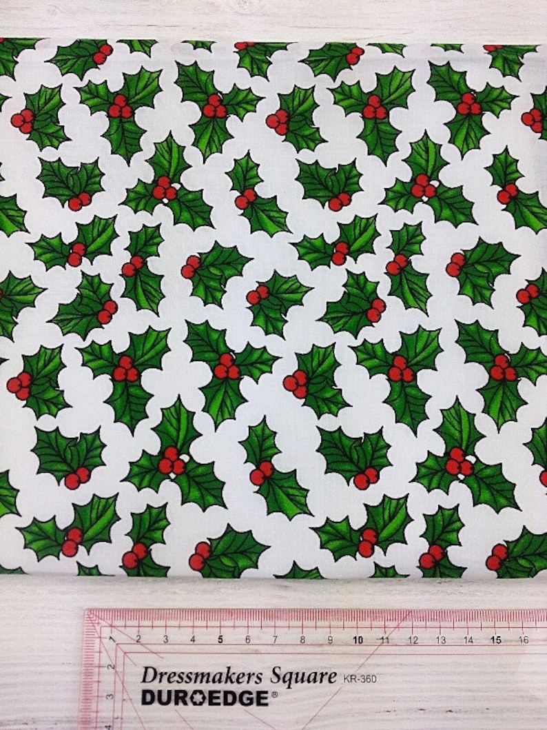 May include: White fabric with a repeating pattern of green holly leaves and red berries. The fabric is labeled "Dressmakers Square" and "DUROEDGE".