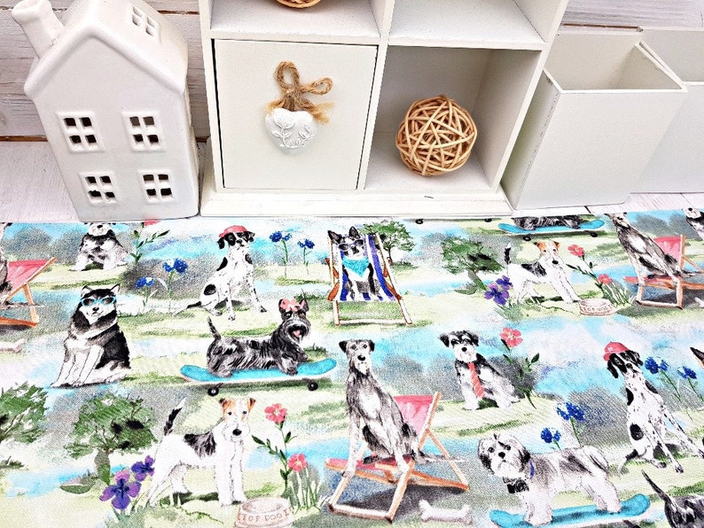 May include: A colorful fabric featuring various dog breeds enjoying leisure activities. Dogs are depicted on skateboards, in beach chairs, and in a park setting with flowers and trees. The fabric has a light blue and green background.