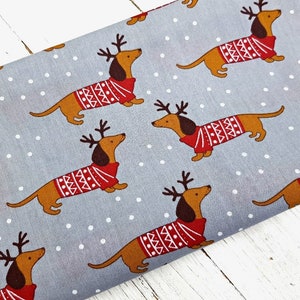 May include: Gray fabric with a repeating pattern of brown dachshunds wearing red and white sweaters with reindeer antlers. The background has white polka dots.