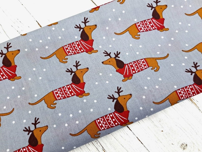 May include: Gray fabric with a repeating pattern of brown dachshunds wearing red and white sweaters with reindeer antlers. The background has white polka dots.