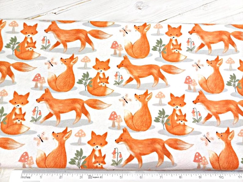 May include: White fabric with a repeating pattern of orange foxes. The foxes are in various poses, including walking, sitting, and holding a smaller fox. There are also small orange mushrooms and butterflies scattered throughout the fabric.