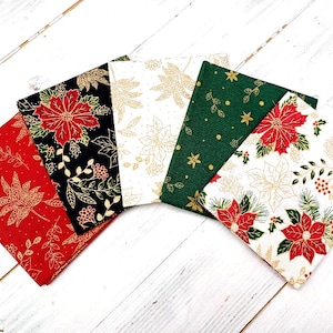 Pode incluir: Cinco quadrados de tecido com tema natalino. Os quadrados apresentam um fundo branco com flores de poinsettia douradas e vermelhas e folhas. Os quadrados estão dispostos em forma de leque.