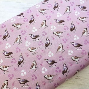 Op de afbeelding: Roze stof met een herhalend patroon van bruine vogels en kleine paarse bladeren. De vogels kijken in verschillende richtingen en bevinden zich in verschillende posities.