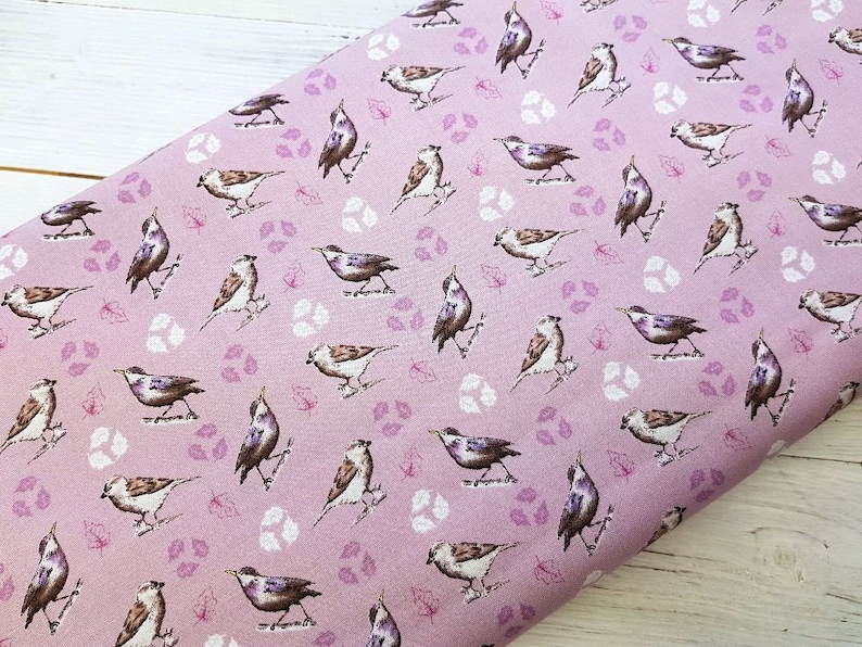 Op de afbeelding: Roze stof met een herhalend patroon van bruine vogels en kleine paarse bladeren. De vogels kijken in verschillende richtingen en bevinden zich in verschillende posities.