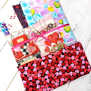 Puede incluir: Una pila de cuadrados de tela con temática de San Valentín. Las telas presentan patrones de corazones, corazones de caramelo con frases como "Kiss" y "Love", y un camión rojo con el texto "Be Mine" y "XOXO". Una cinta rosa envuelve la pila.
