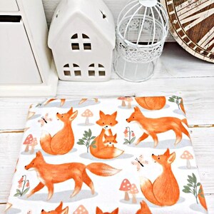 May include: White fabric with a repeating pattern of orange foxes and mushrooms. The foxes are in various poses, including sitting, standing, and walking. Some foxes have a smaller fox cub with them. There are also small butterflies and green leaves in the pattern.