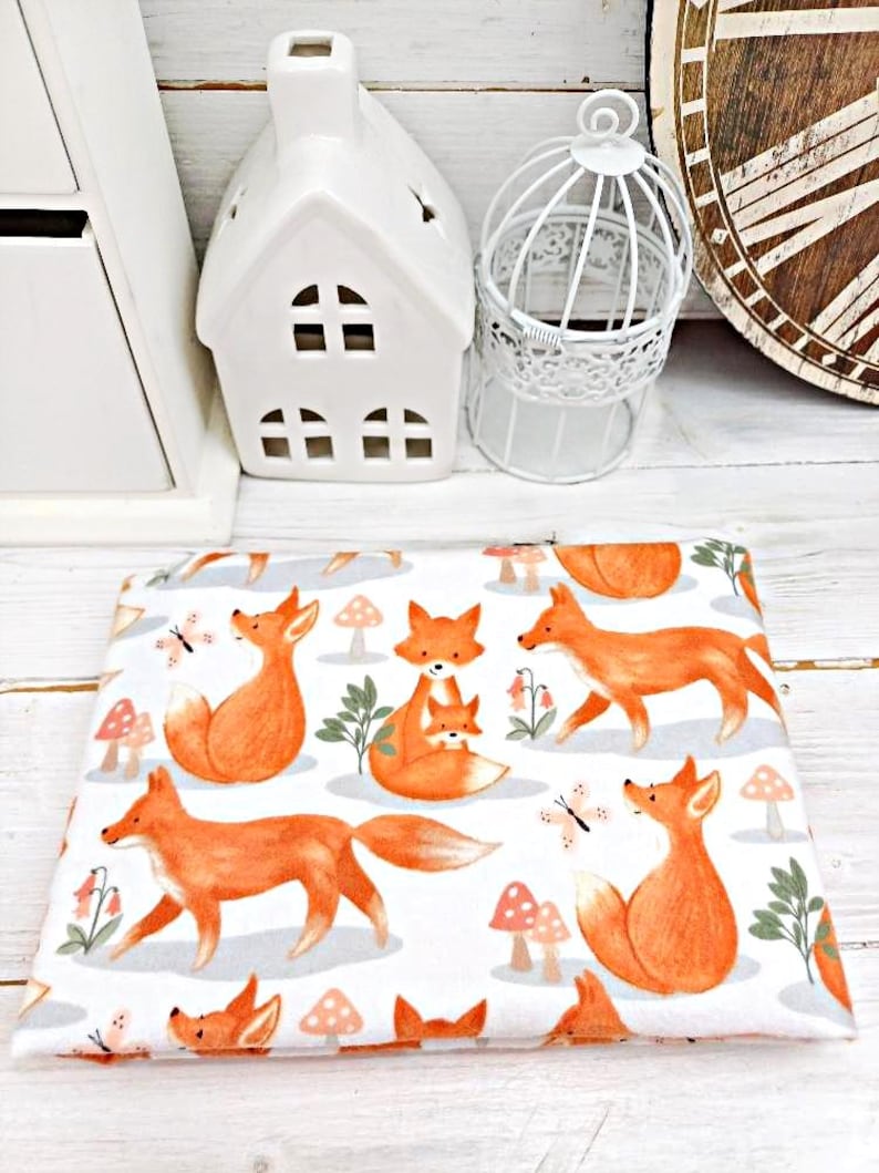 May include: White fabric with a repeating pattern of orange foxes and mushrooms. The foxes are in various poses, including sitting, standing, and walking. Some foxes have a smaller fox cub with them. There are also small butterflies and green leaves in the pattern.