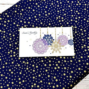 May include: A white greeting card with the text "Season's Greetings" and snowflake designs in purple, gold, and blue. The card is set against a navy blue fabric with a pattern of yellow stars.