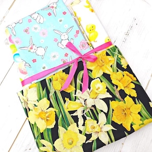 May include: A bundle of three fabric squares with different patterns. The top square is blue with white bunnies and pink flowers. The middle square is white with a yellow chick. The bottom square is black with yellow daffodils. The squares are tied together with a pink ribbon.