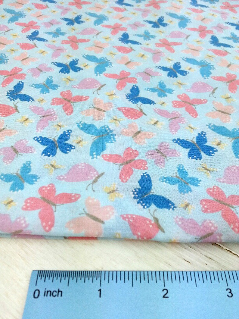 May include: A light blue fabric with a repeating pattern of pink, blue, and yellow butterflies. A ruler is included to show the scale of the fabric.