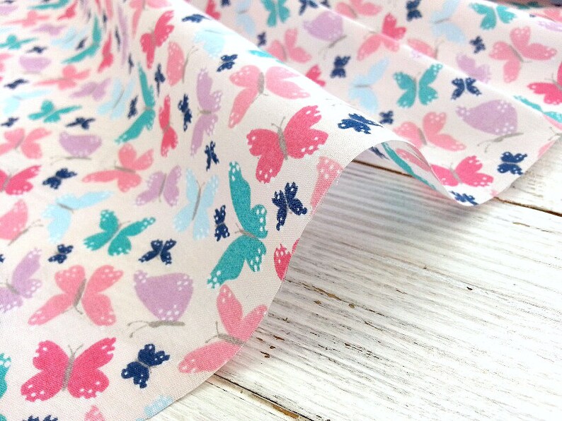 May include: White fabric with a repeating pattern of colorful butterflies. The butterflies are in shades of pink, blue, teal, and purple.
